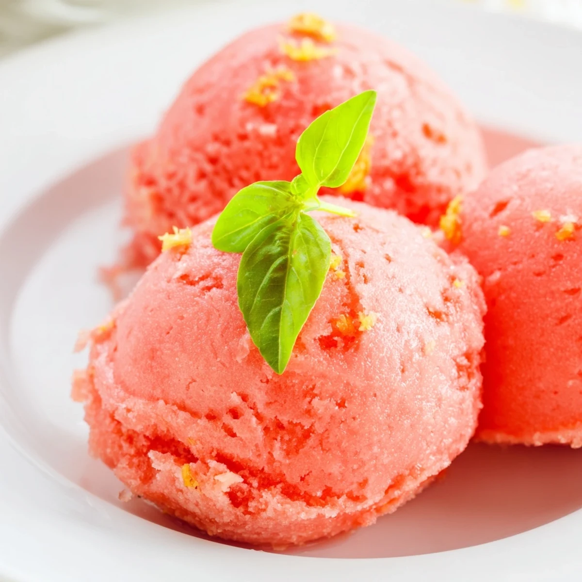 Pale pink sorbet with basil garnish sits beside sliced grapefruit on a rustic wooden table.