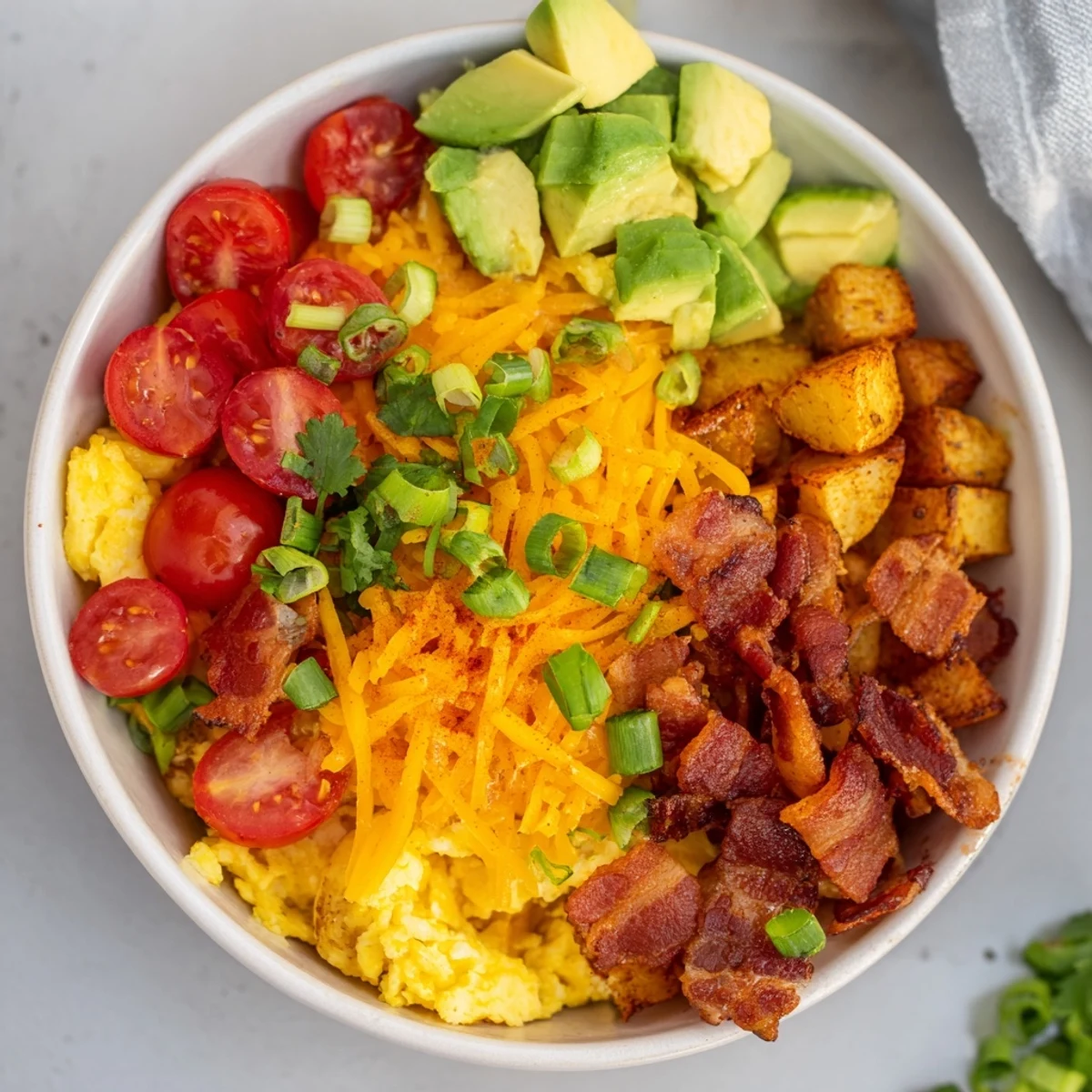 A vibrant Loaded Breakfast Bowl featuring golden roasted potatoes, scrambled eggs, crispy bacon, and melted cheddar cheese topped with fresh avocado and tomatoes.