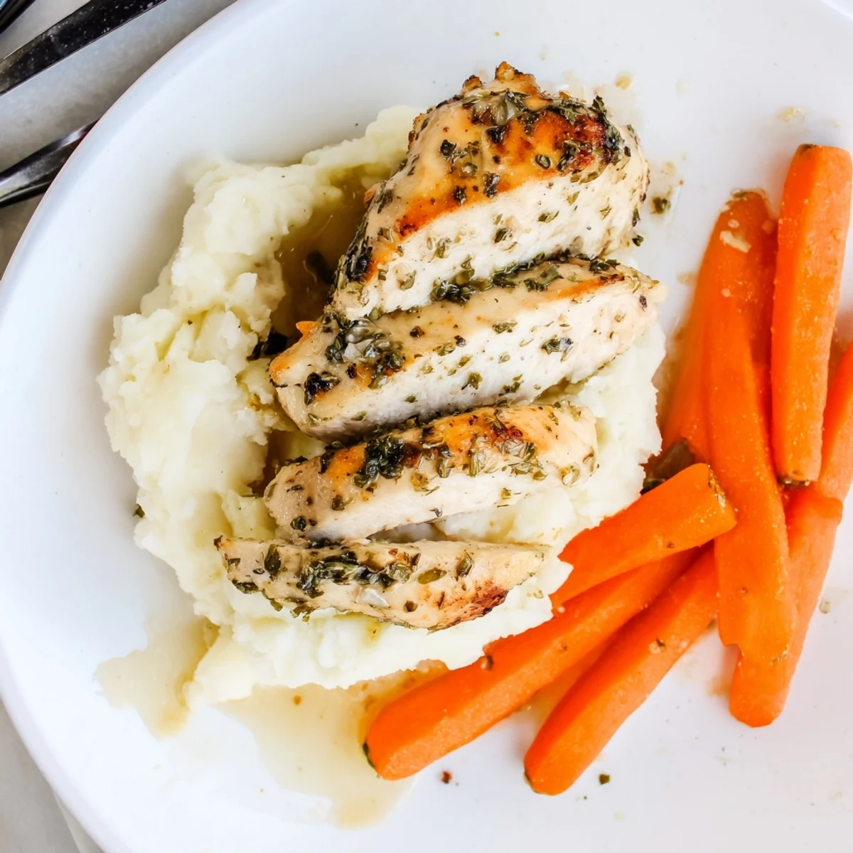 Golden herb-crusted chicken breast resting beside fluffy mashed potatoes and honey-glazed carrot sticks on a white plate